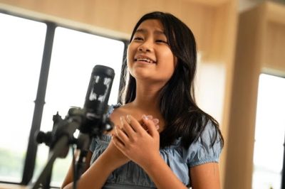 Close-up of a smiling young girl singing with emotion with her hands over her chest in front of a studio condenser microphone