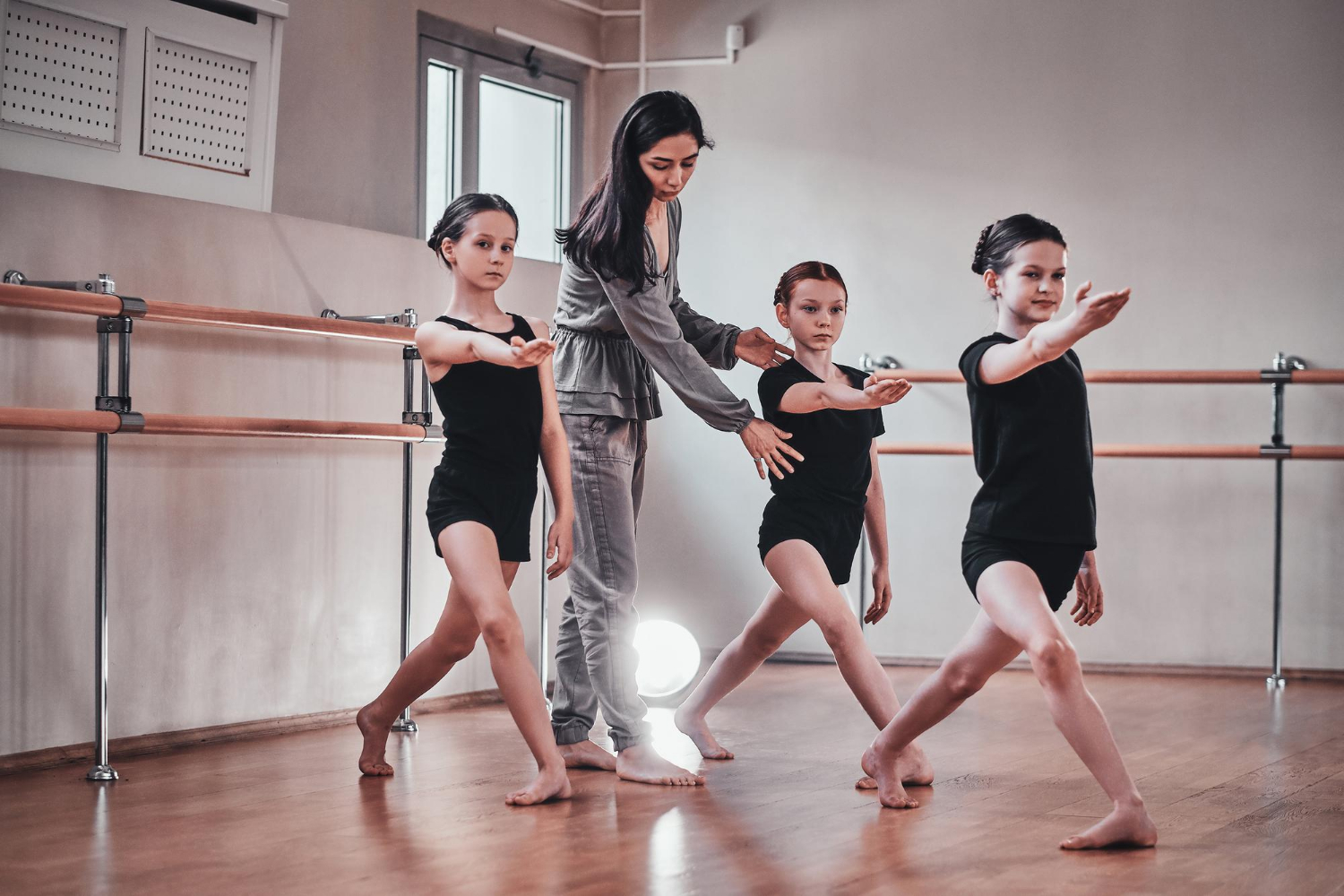 A ballet instructor adjusts the posture of three young students practicing a dance step in a studio with wall barres