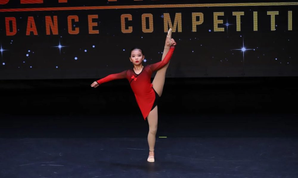 Young girl in a red and black dance costume performing a high vertical leg extension on stage during a dance competition