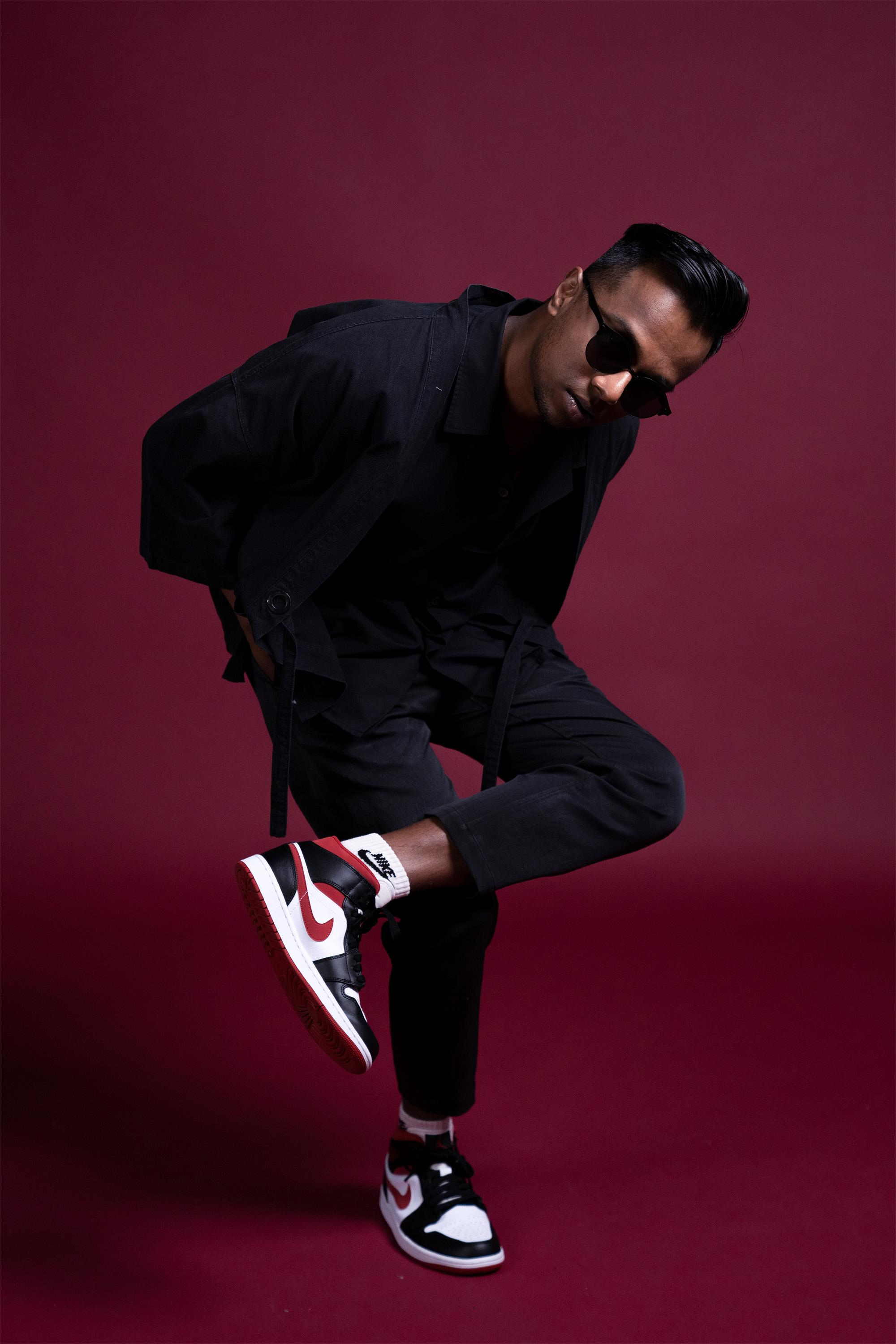 male-dancer-posing-in-black-streetwear-studio-portrait A man wearing sunglasses, an all-black streetwear outfit, and nike shoes while striking a pose