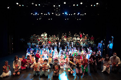 Young performers holding flowers posing together on stage with a seated theater audience in the background