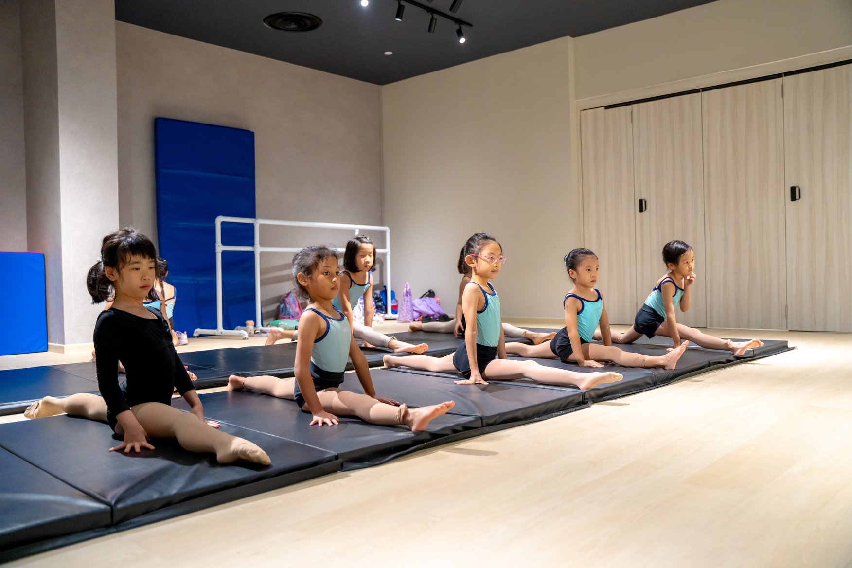 Row of young dancers lined up on floor mats holding side splits during flexibility practice