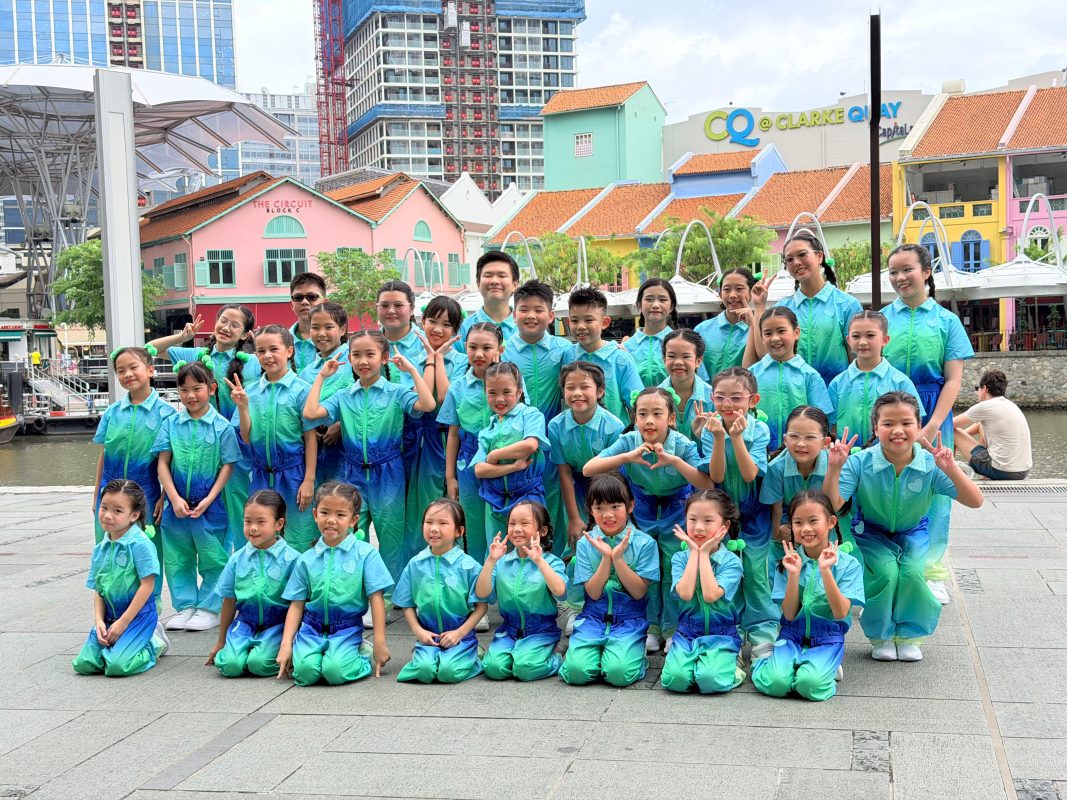 A large group of children wearing matching blue and green gradient outfits pose together outdoors with colorful buildings in the background