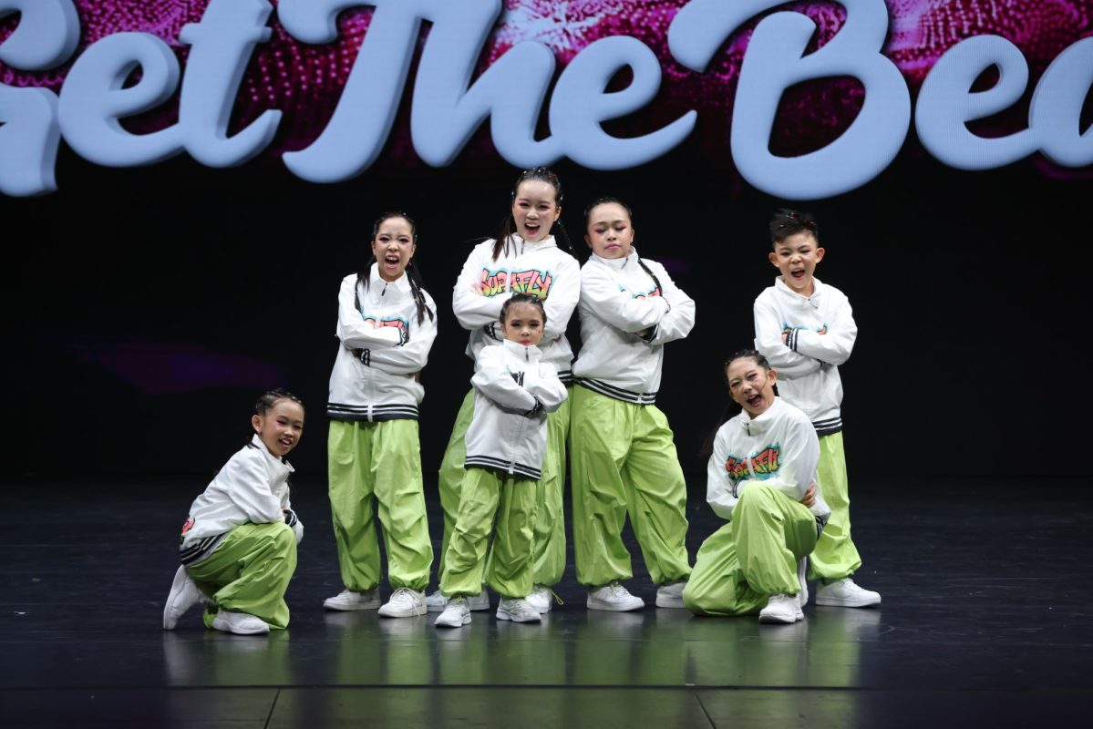 A group of young dancers wearing white zip-up jackets and light green track pants pose together during a hip-hop dance competition stage performance
