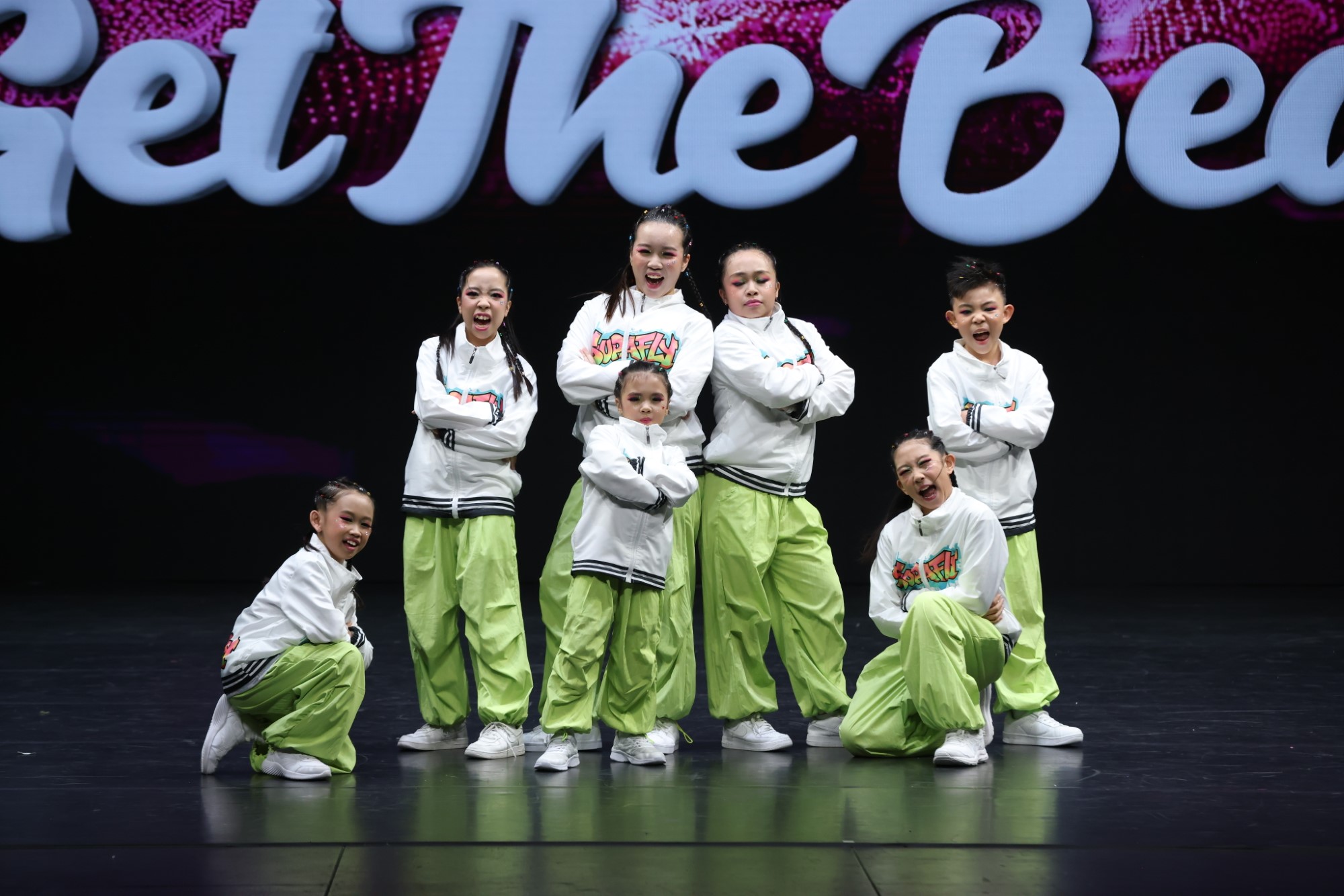 A group of young dancers wearing white zip-up jackets and light green track pants pose together during a hip-hop dance competition stage performance