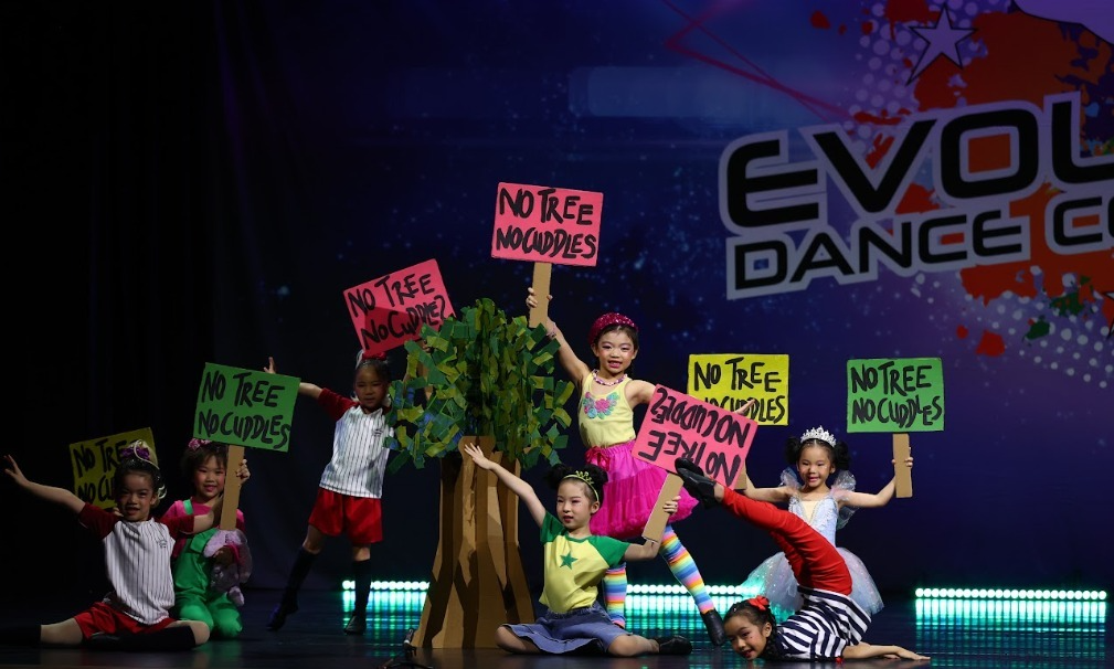 Group of young children performing a dance routine on stage around a prop tree holding signs that read No Tree No Cuddles at Evolution Dance Competition